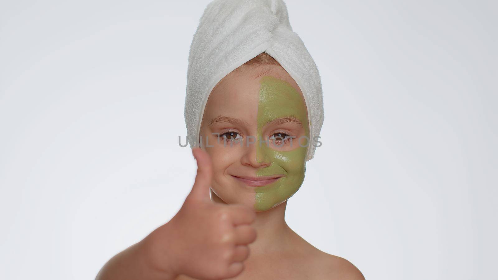 Beautiful young smiling child girl in towel on head applying cleansing moisturizing green mask on face, showing thumbs up. Teenager kid face skin care treatment, natural cosmetics. Female portrait