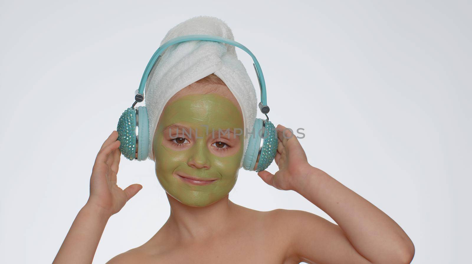 Smiling young child girl after bath in towel on head and moisturizing green mask on face listening to music on headphones, dancing. Teenager kid skin care treatment, natural cosmetics. Female portrait