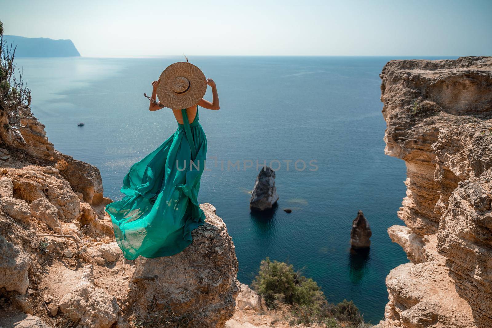 A girl with loose hair in a long mint dress descends the stairs between the yellow rocks overlooking the sea. A rock can be seen in the sea. Sunny path on the sea from the rising sun.