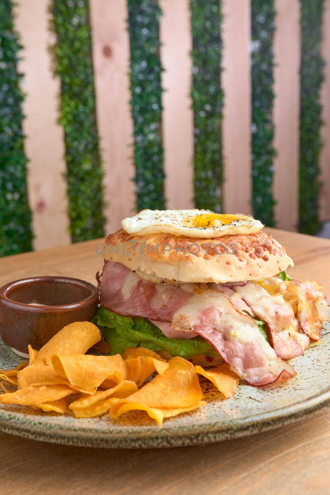 photo of delicious bacon burger accompanied with an egg, chipotle dressing and dehydrated bananas on wooden table. big hamburguer.