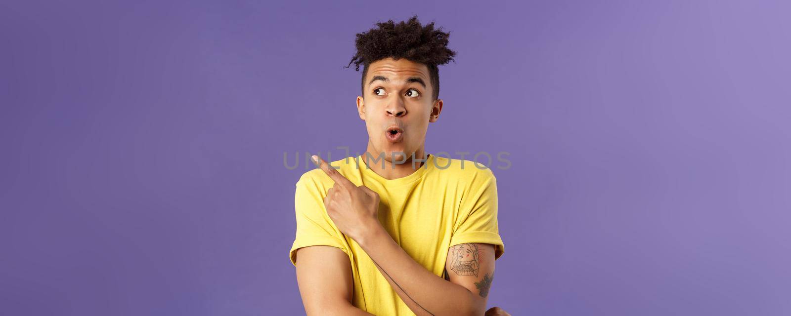 Waist-up portrait of amazed, impressed hispanic male with dreads, seeing something incredible and cool, pointing finger looking upper left corner, breathtaking awesome thing, purple background.