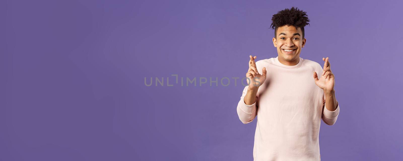 Portrait of hopeful positive, smiling young man having faith in dreams may come true, make wish, cross fingers good luck and awaiting for miracle, standing purple background praying by Benzoix