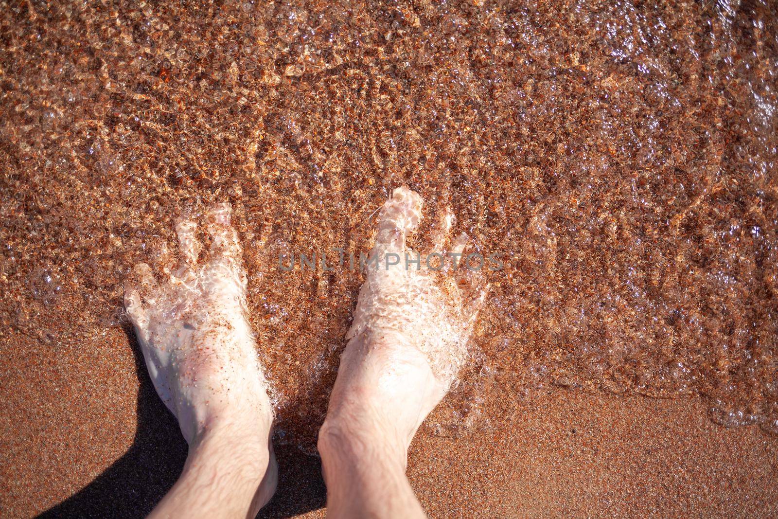 Top view of men's legs in the sea. A man is standing on the sand. An ocean wave washes the shore. Summer holidays.