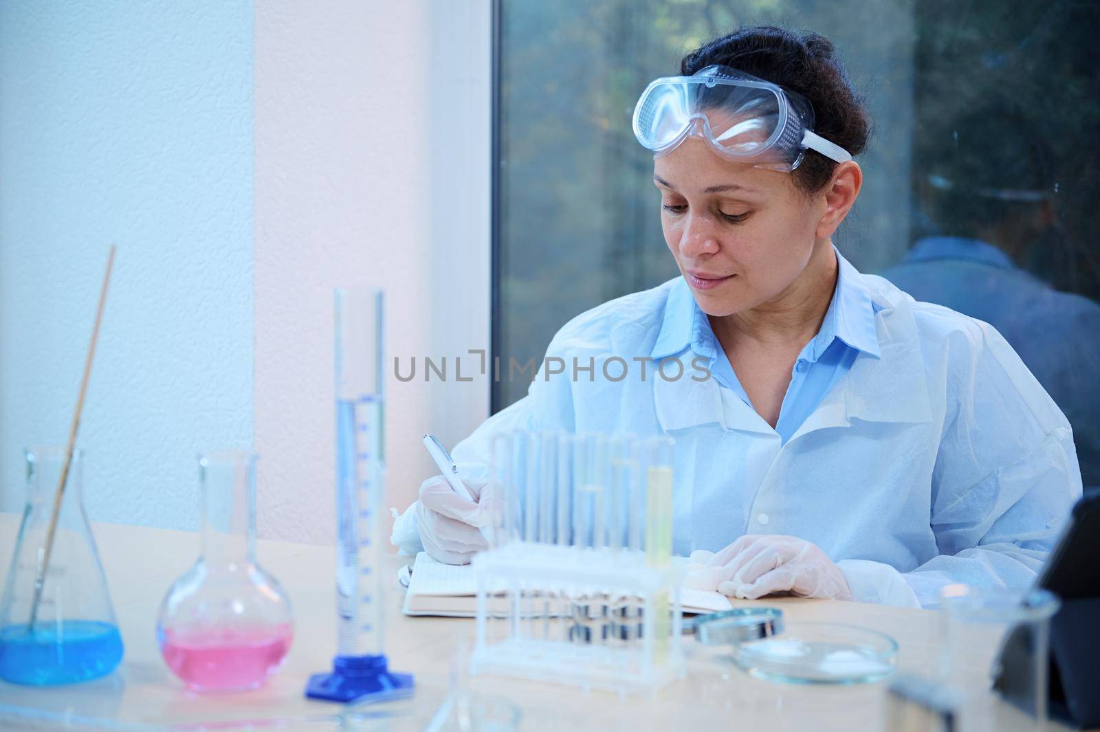 Confident multi-ethnic woman scientist, chemist-pharmacologist sits at her desk with lab glasswear and writes down the results of experiments carried out in a pharmacological laboratory in a notepad