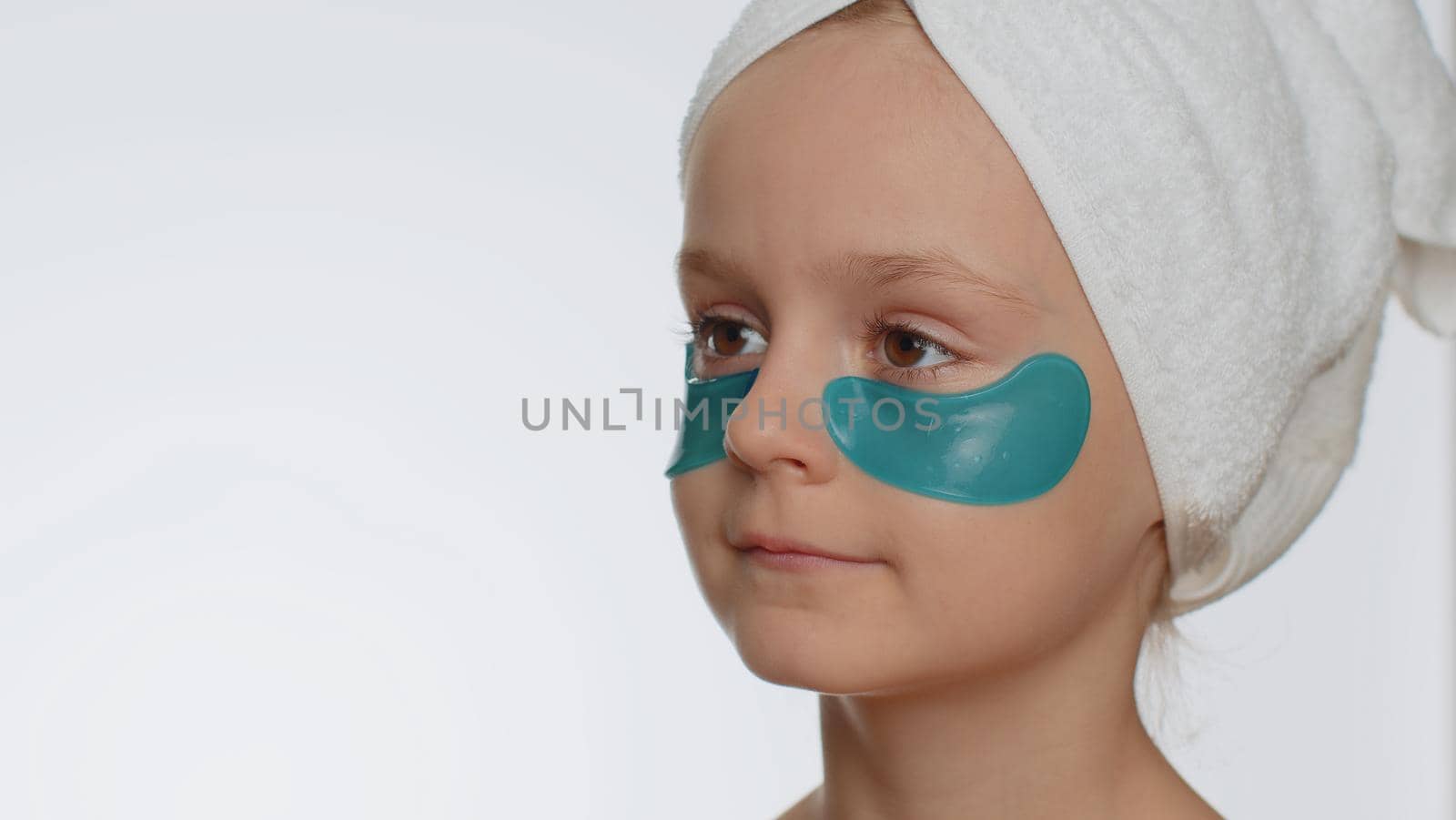 Beautiful young smiling child girl in towel on head applying blue patches under eyes. Teenager kid face skin care treatment, natural cosmetics. Female portrait on white background. Perfect fresh clean