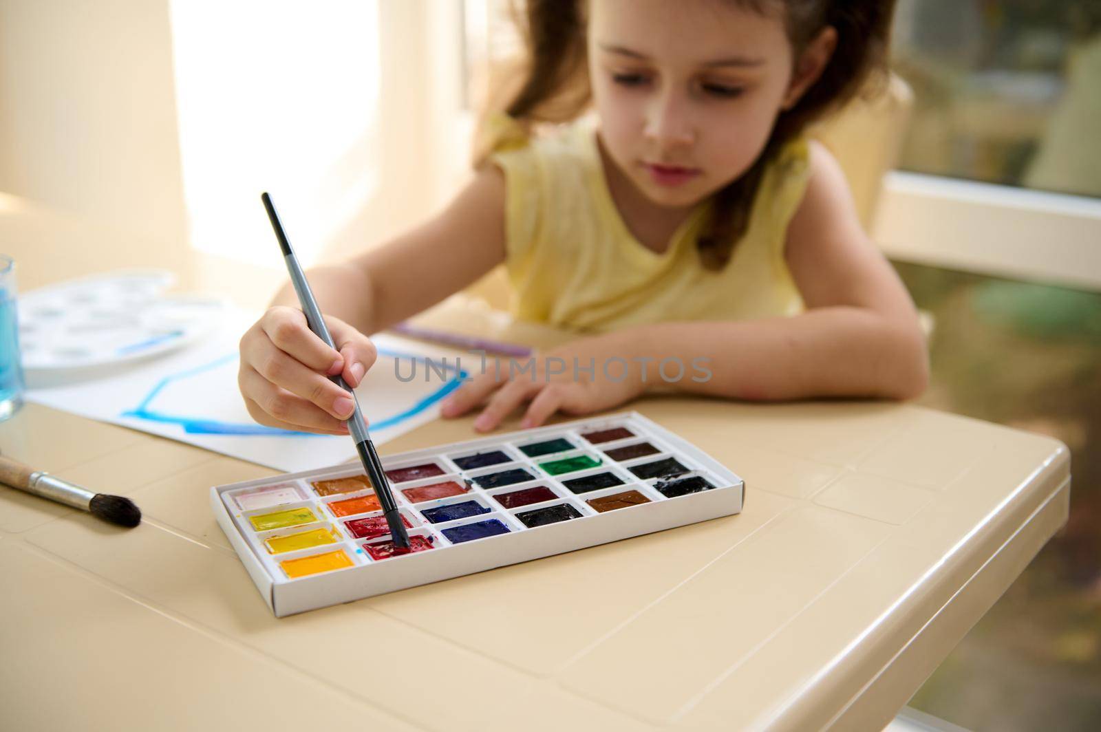 Focus on the hand of a talented little girl, holding paintbrush and mixing colorful paints on a professional watercolor palette. Learning peinture. Art class. Kids education and hobby. Back to school
