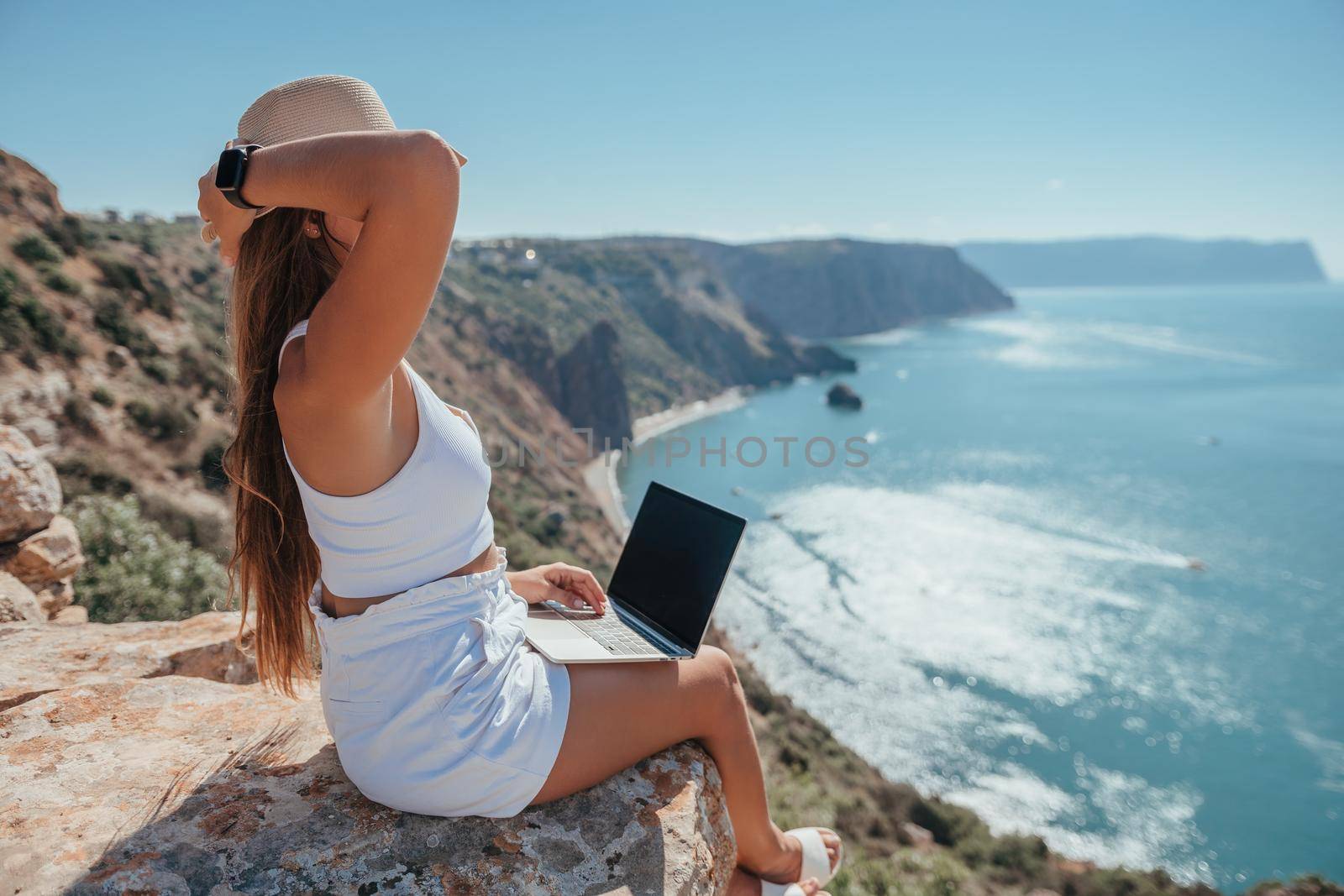 Successful business woman in yellow hat working on laptop by the sea. Pretty lady typing on computer at summer day outdoors. Freelance, travel and holidays concept.