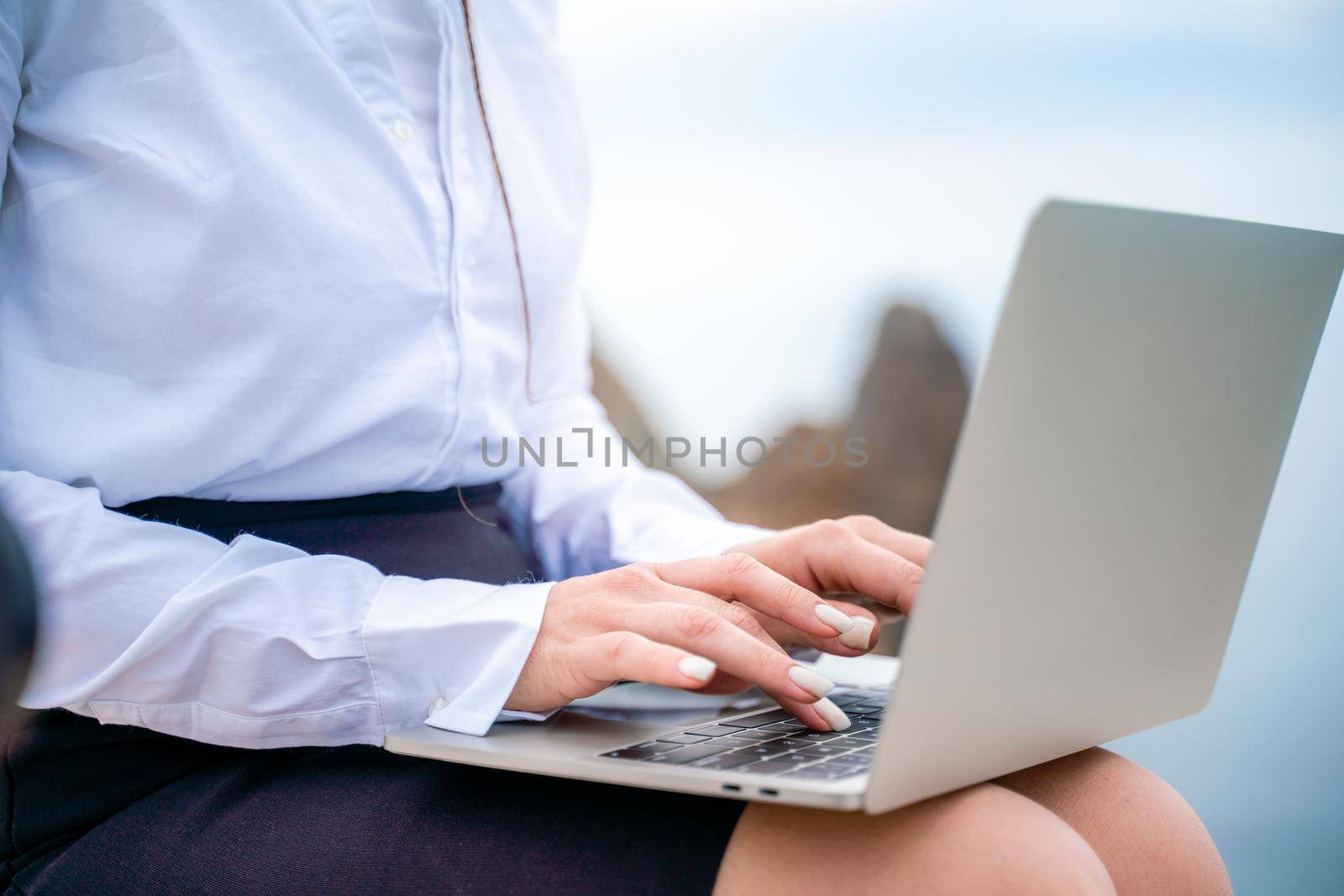 A woman is sitting and typing on a macbook keyboard on a terrace with a beautiful sea view. Wearing a white blouse and black skirt. Freelance travel and vacation concept, digital nomad