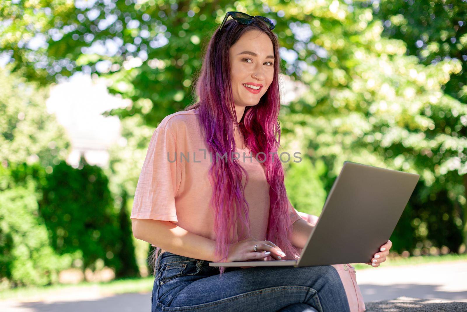 Young girl with pink hair is studying in the spring park, sitting on the wooden bench and browsing on her laptop