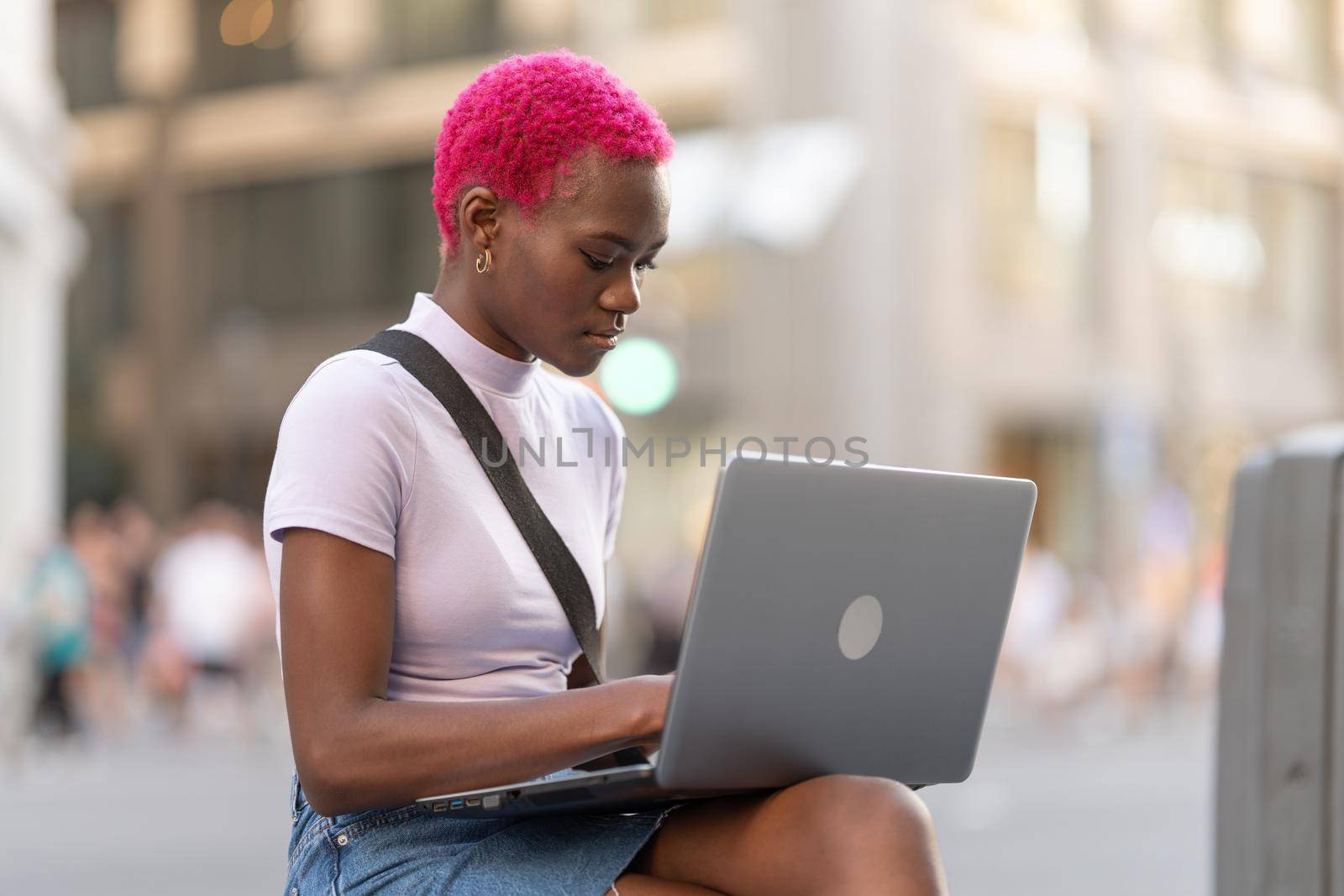 Portrait of an african young woman with pink short hair working with laptop sitting on the street
