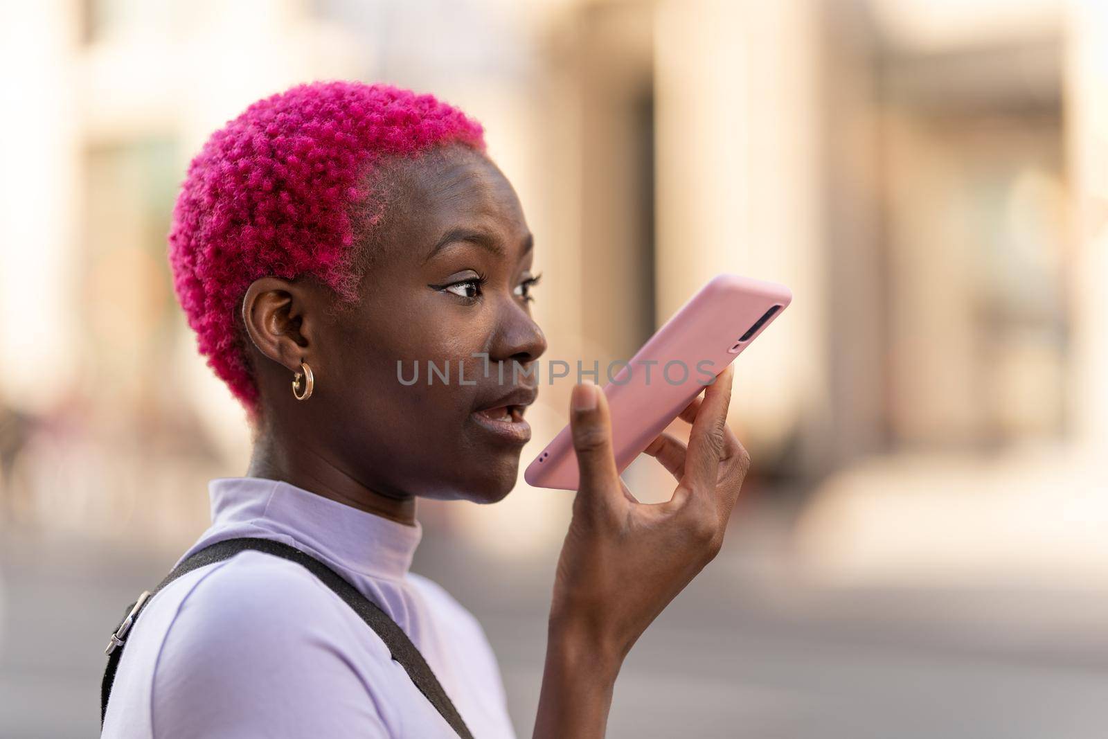 Profile of an afro young woman with pink hair sending a voice message with the mobile outdoors