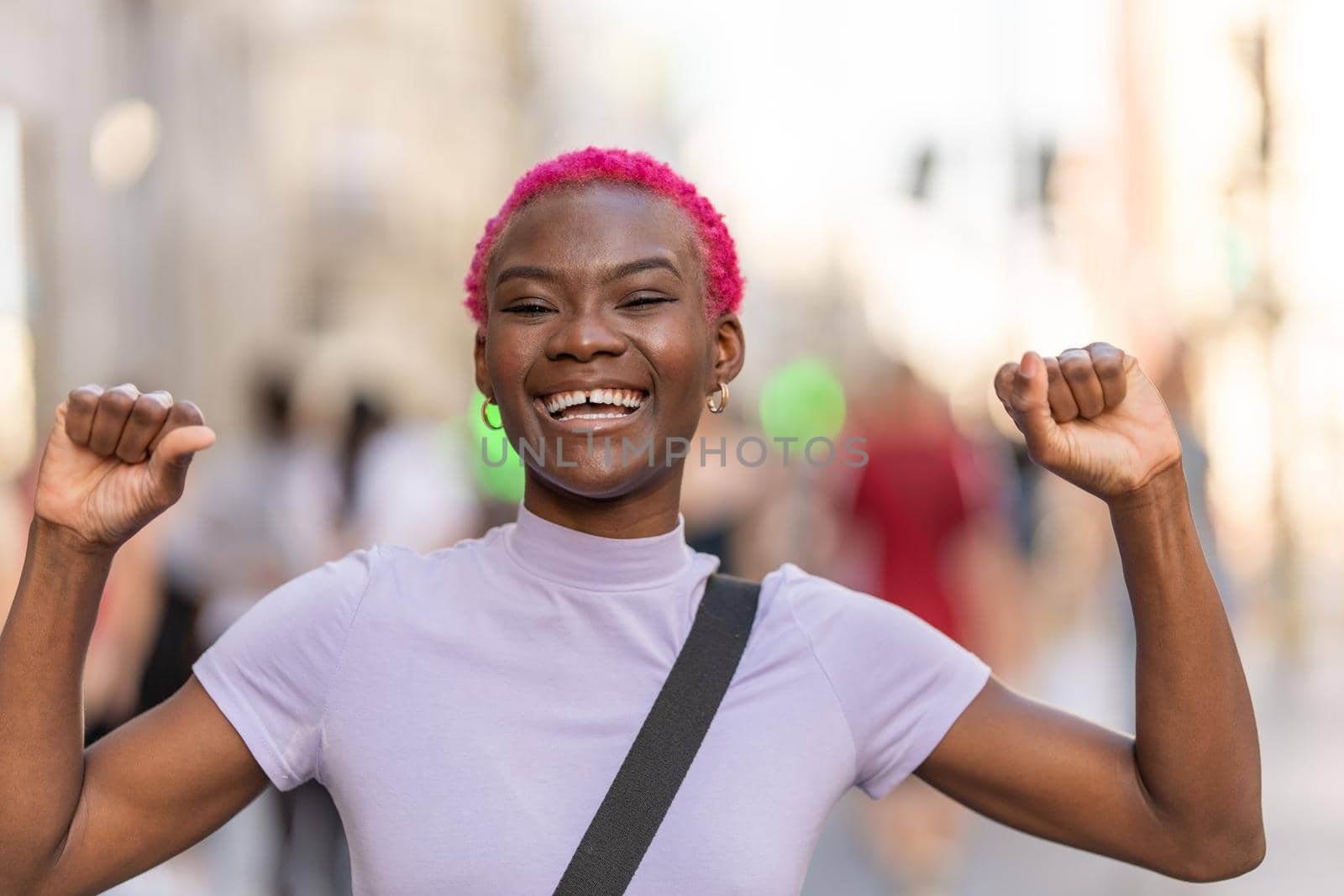 Happy young stylish afro woman smiling to the camera while raising the arms in the city street