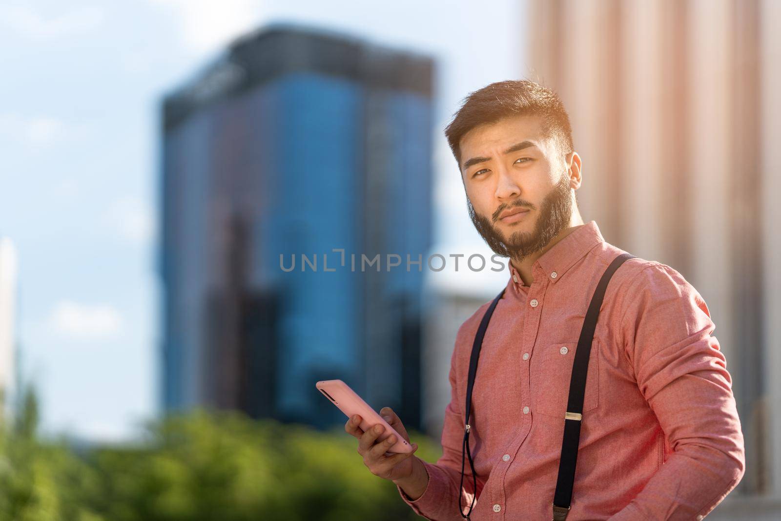 Asian businessman in a shirt and braces looking at the camera while using a mobile outdoors