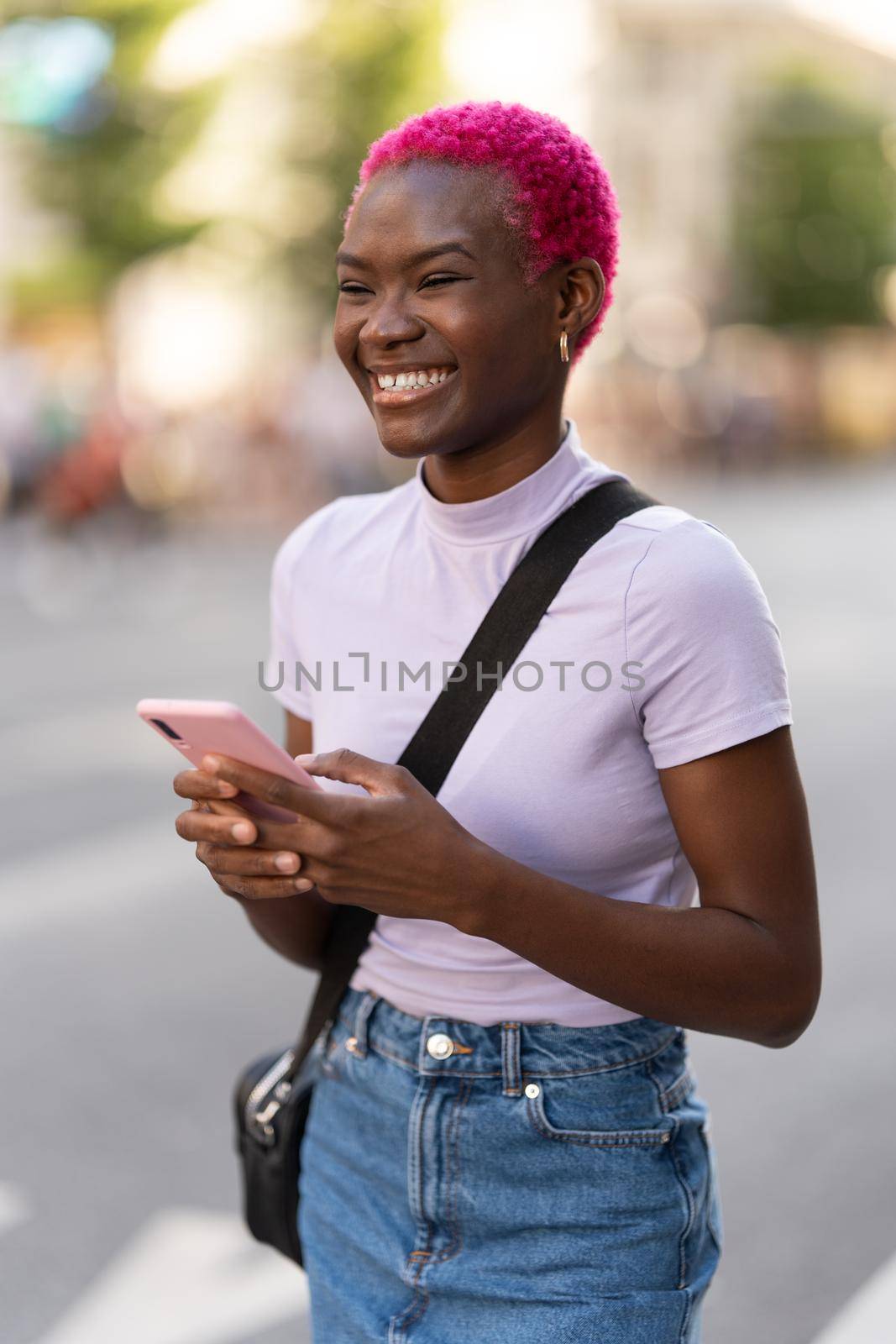 African young woman smiling while using the mobile outdoors by ivanmoreno