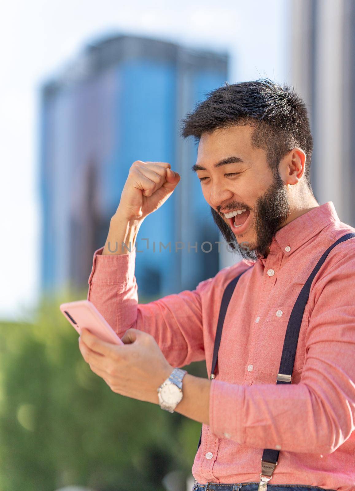 Vertical photo of an asian businessman gesturing success while using to the mobile outdoors