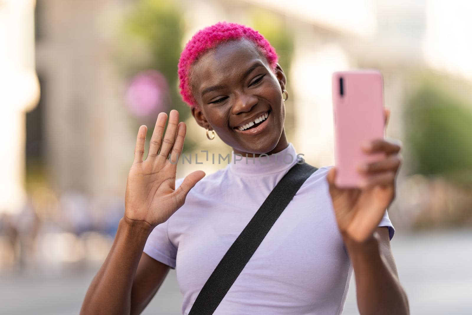 African young woman with pink short hair gesturing hello to the mobile in the street
