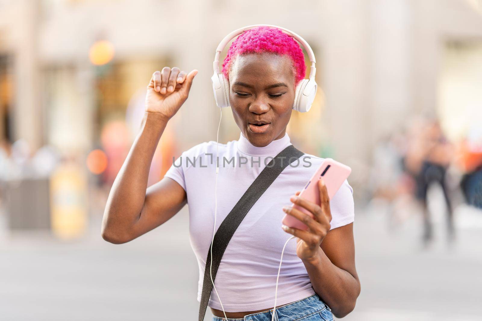 Stylish afro woman dancing while listening to music with headphones and a mobile in the street