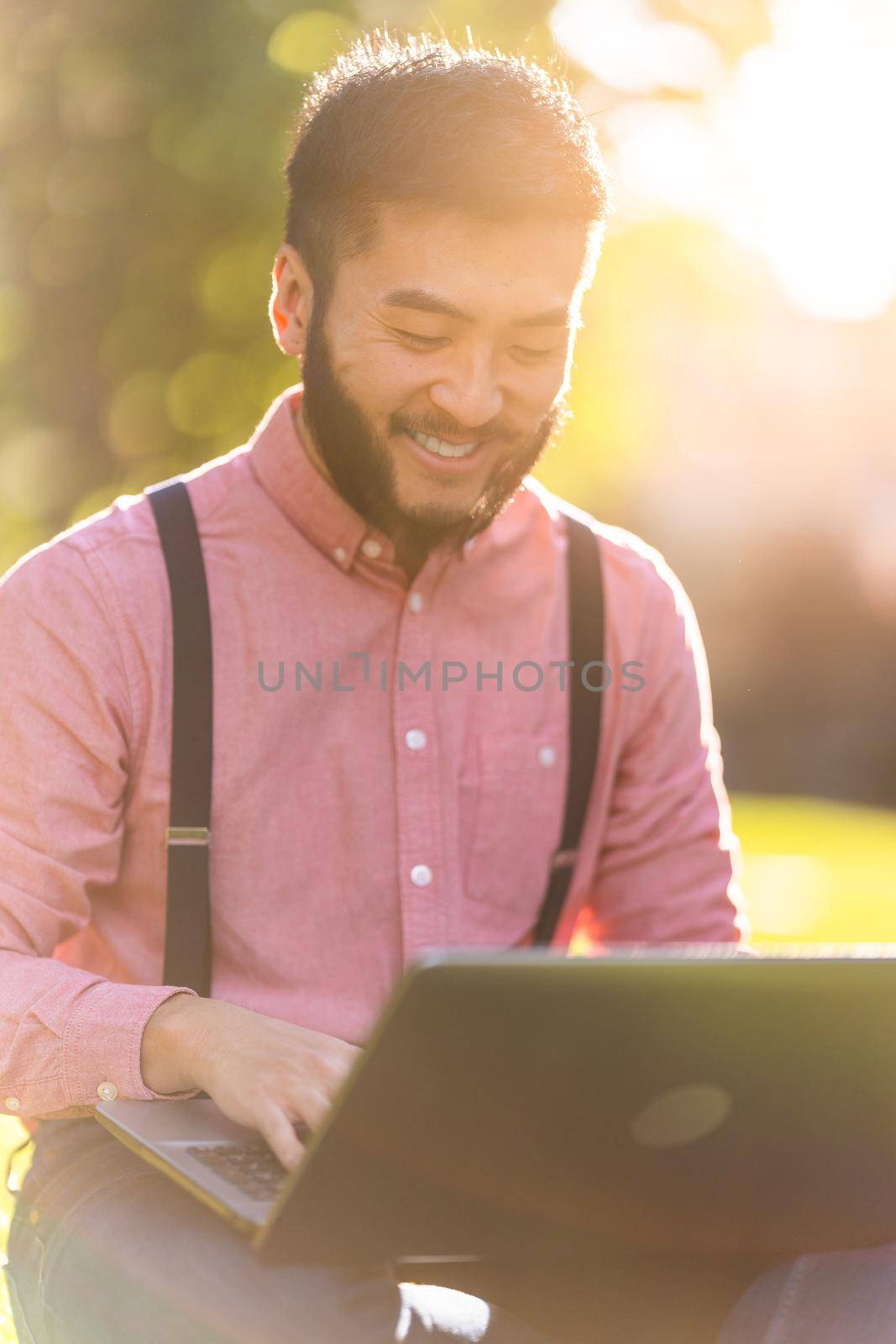 Sunlight illuminating a chinese man in a shirt and braces working on a laptop in an urban park