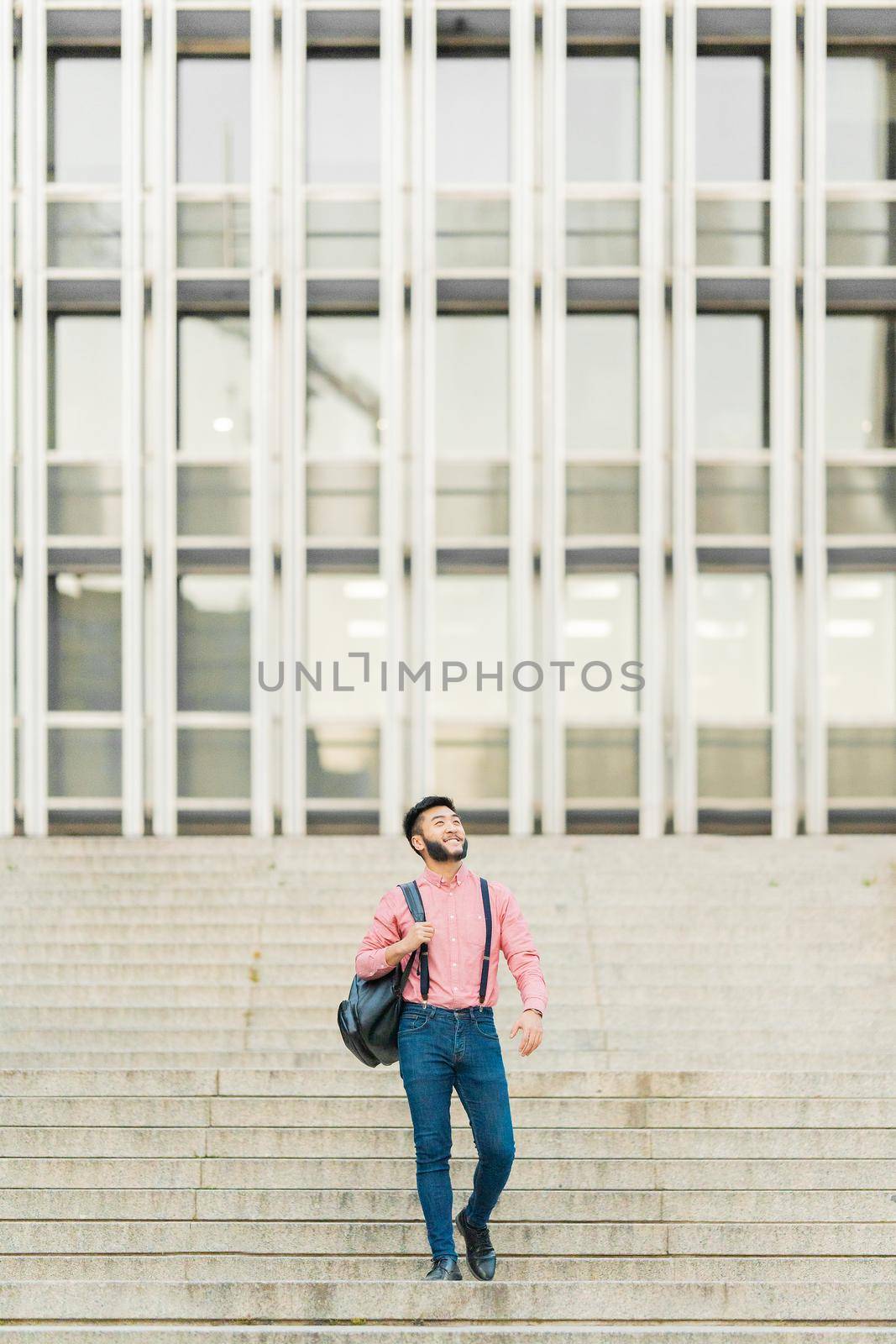 Asian man in casual clothes walking down a stairs with a business building in the background