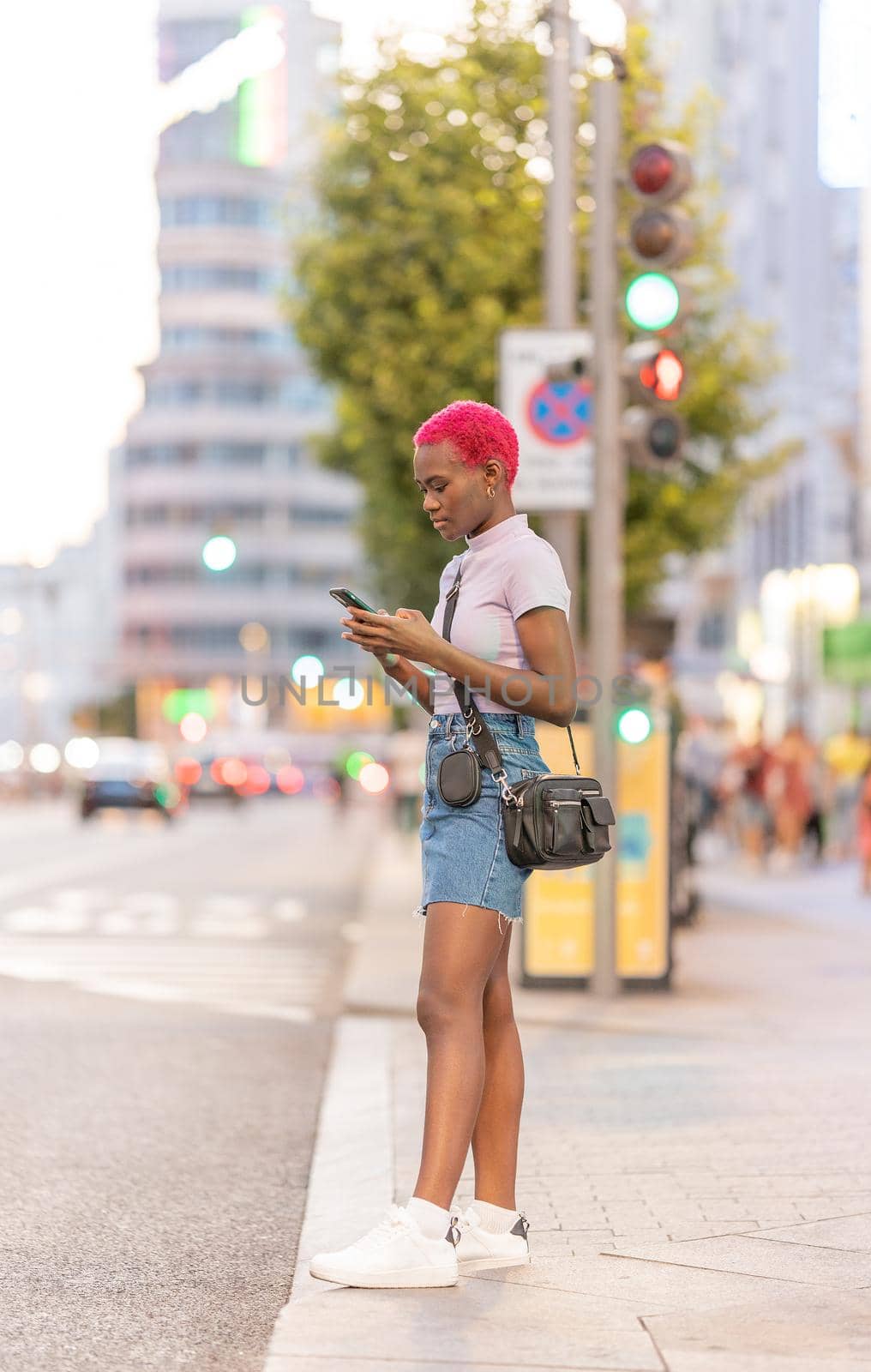 Vertical photo of an afro young woman standing on the street using the mobile during sunset