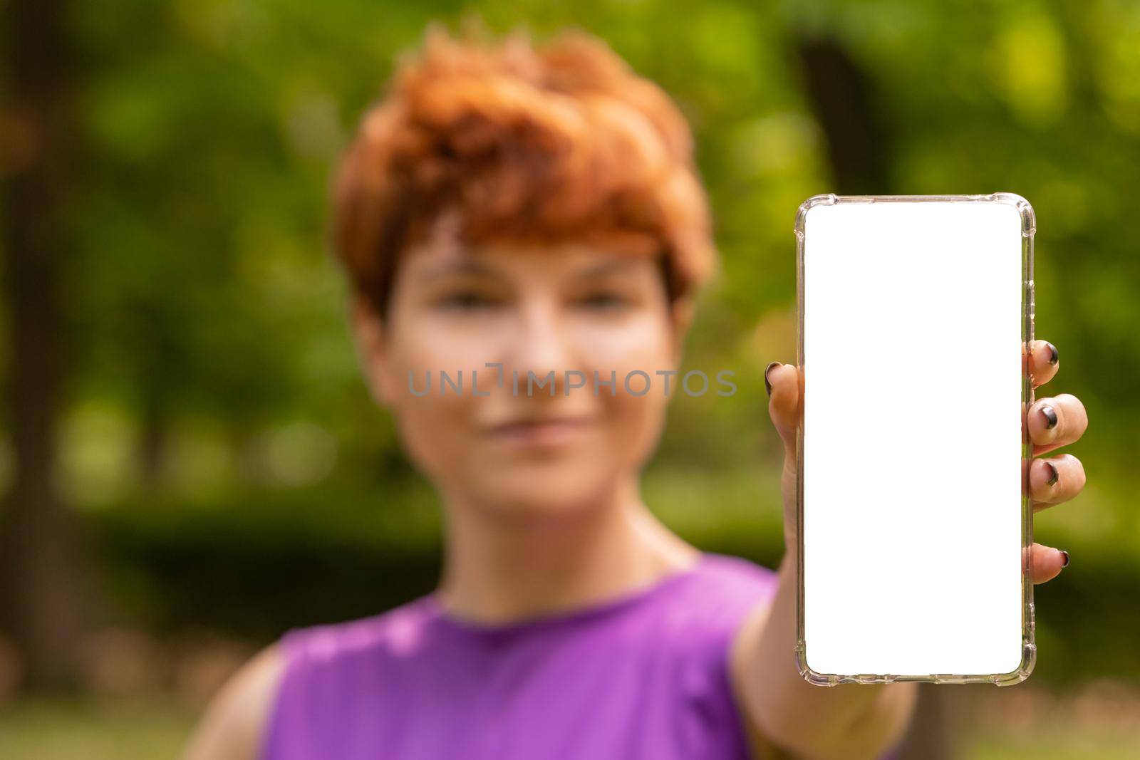 Soft focus of androgynous female demonstrating cellphone with blank screen while advertising app on summer day in park