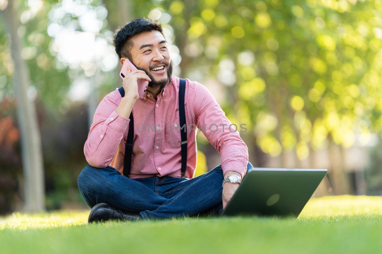 Asian man in shirt and braces smiling while talking on a mobile phone and working on a laptop sitting in a park