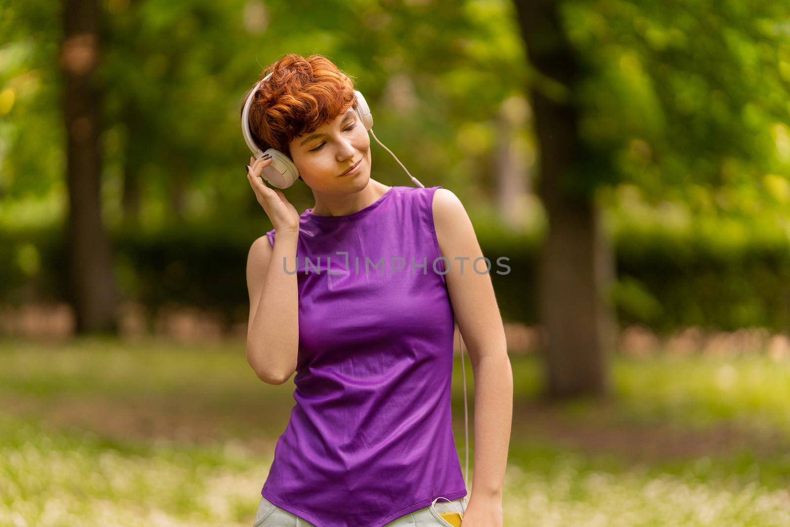 Non binary meloman in casual clothes with short red hair holding hand in pocket, and adjusting headset while listening to music with closed eyes on blurred background of park on weekend