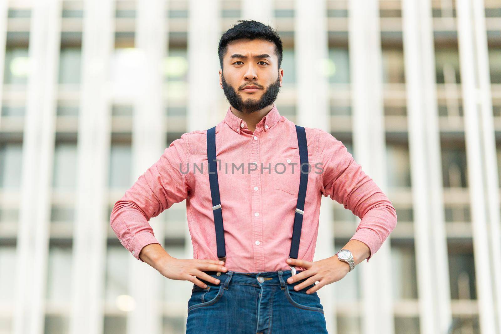 Serious asian man in casual clothes posing with his arms on his hips looking at the camera outdoors