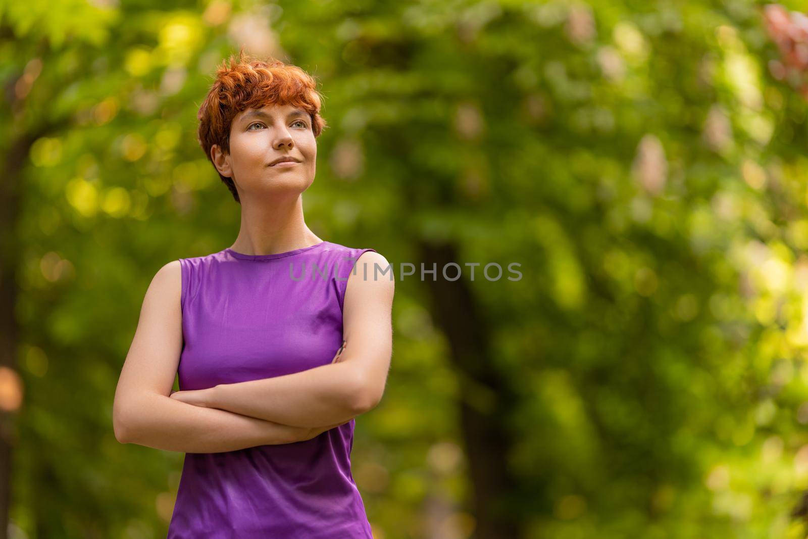 Self assured androgynous redhead female in purple top crossing arms and looking away on blurred background of green trees on summer weekend day in garden