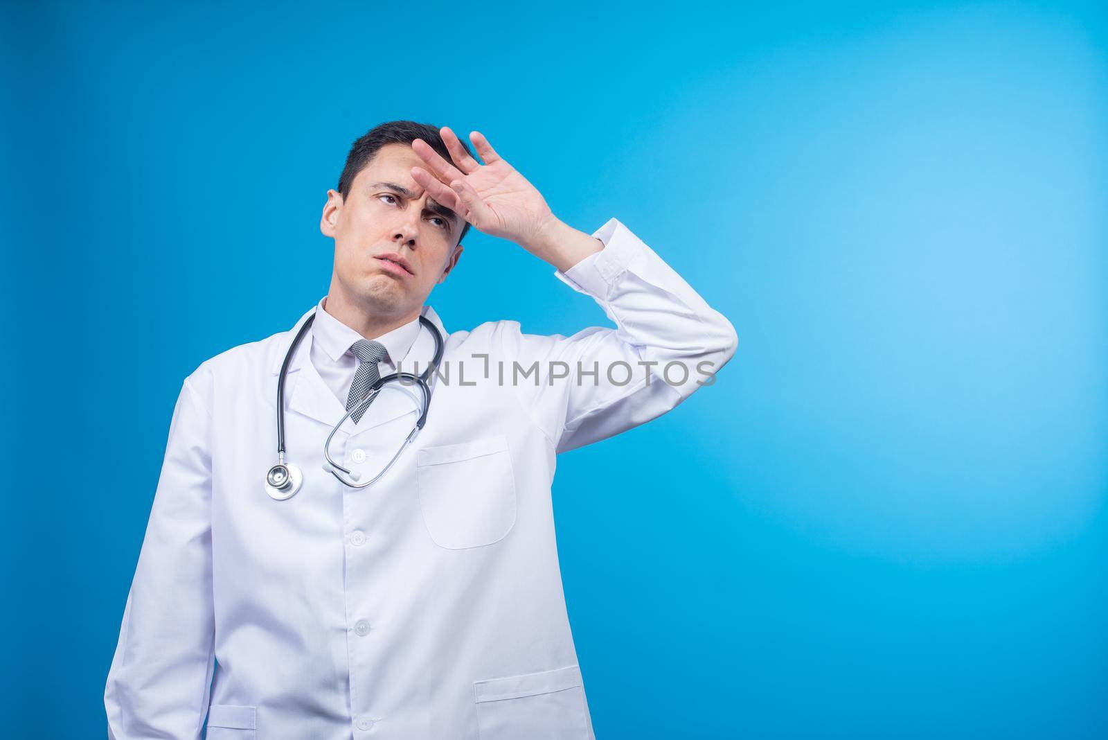 Physician with stethoscope touching forehead and looking away while being tired at work on blue backdrop