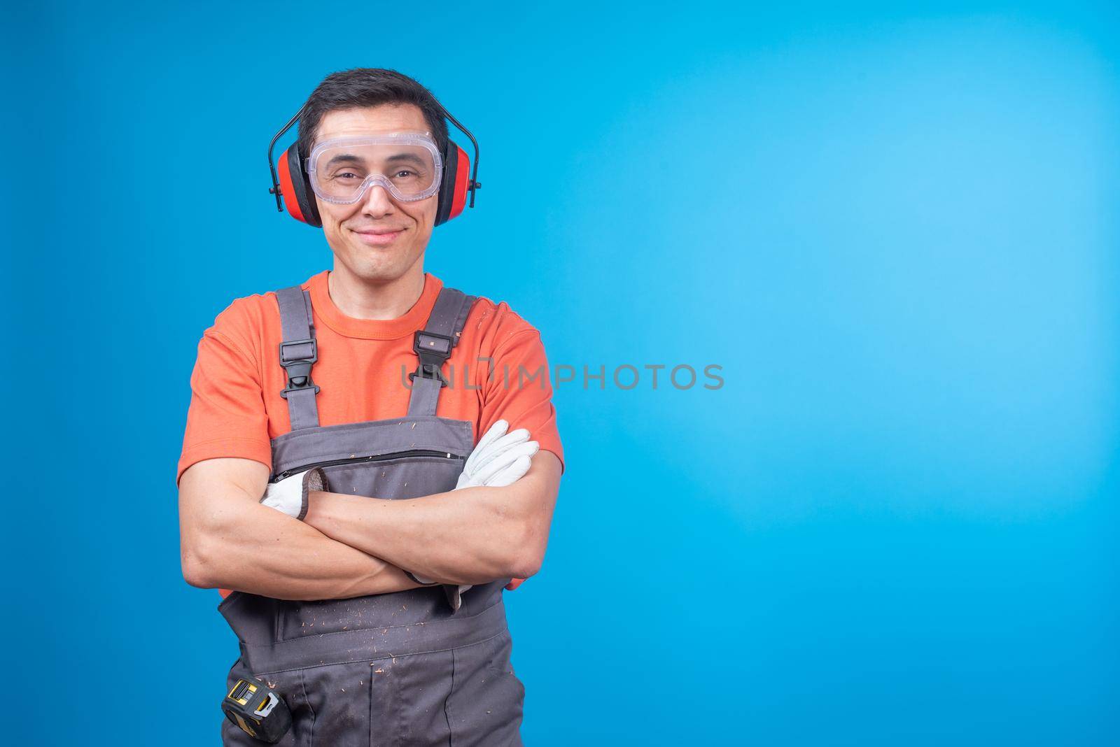 Positive male carpenter in uniform with protective goggles and earmuffs crossing arms and looking at camera with smile against blue background