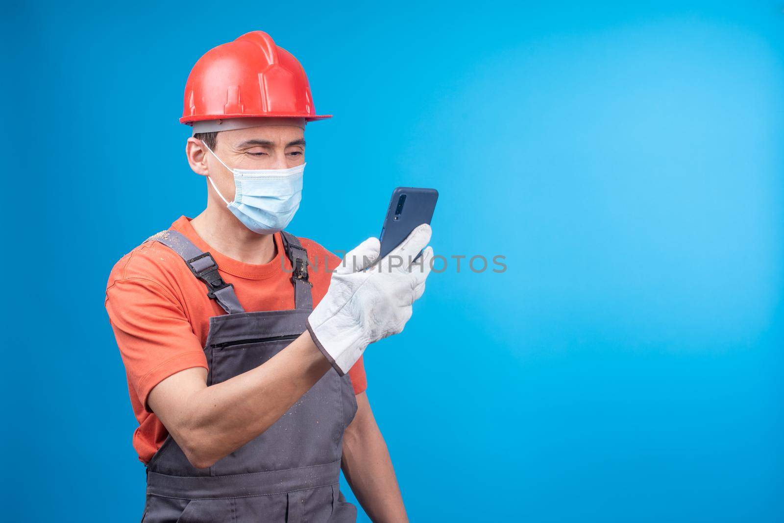 Man in workwear with gloves, face mask and hardhat watching video on mobile phone while taking break in work against blue background during pandemic