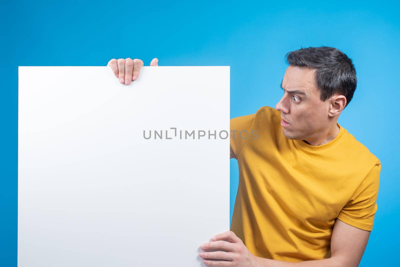 Young startled man in yellow t shirt looking at white paper sheet in hands on blue background