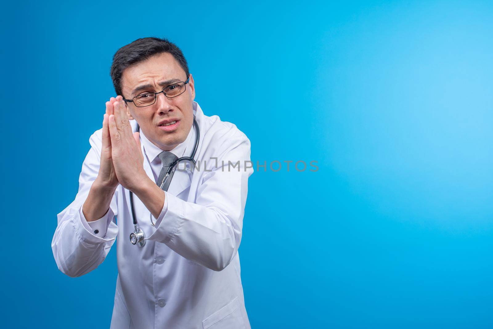 Young male doctor in glasses and medical uniform looking at camera imploringly while standing with folded hands against blue backdrop