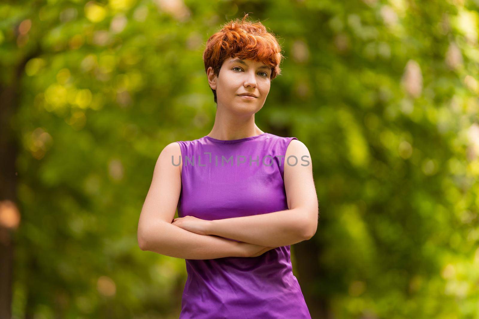 Confident person of non binary gender identity crossing arms, and looking at camera while standing on blurred background of lush trees on summer day in park