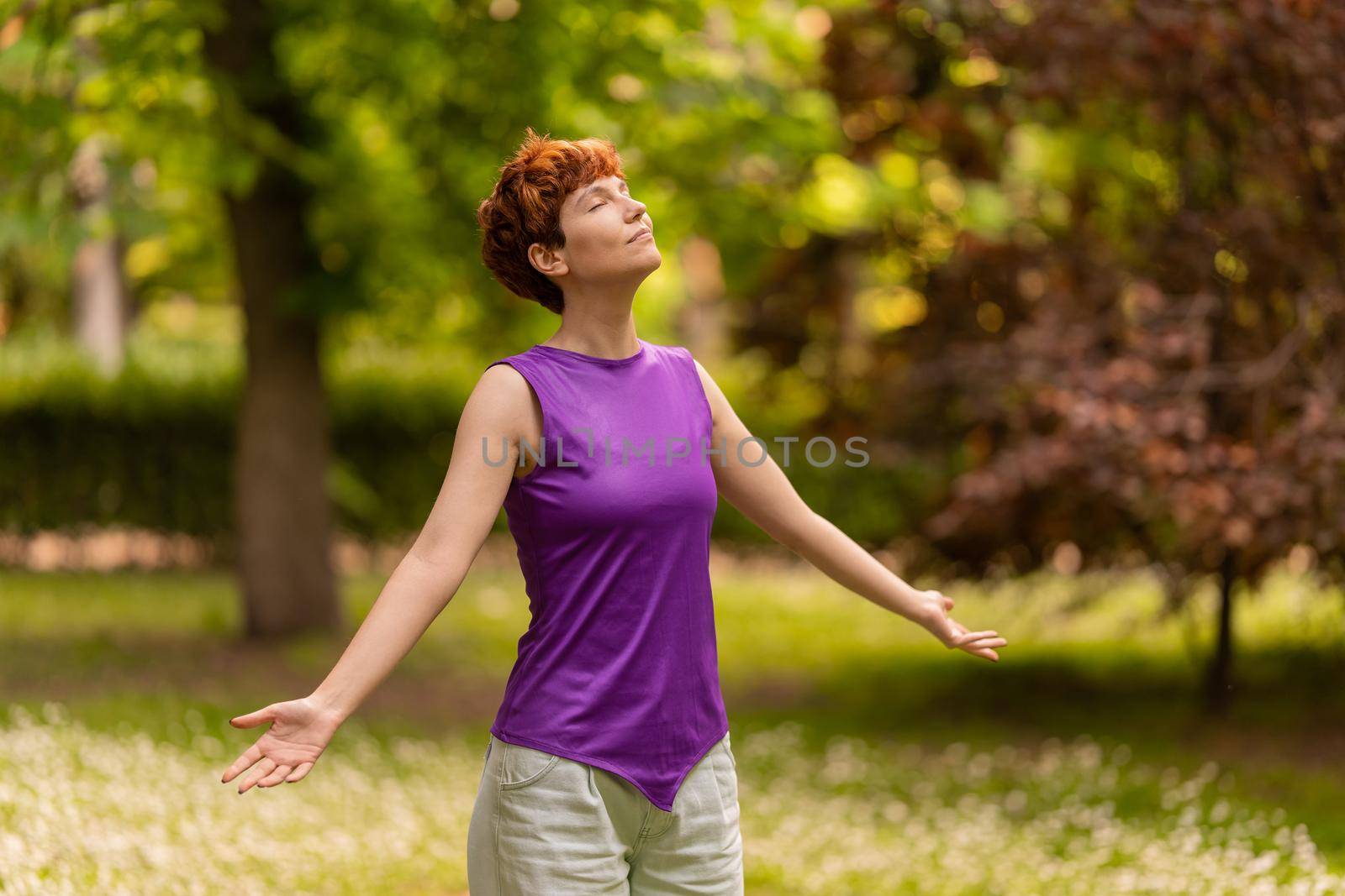 Calm lesbian female in casual clothes spreading arms and closing eyes while breathing fresh air on weekend day in park