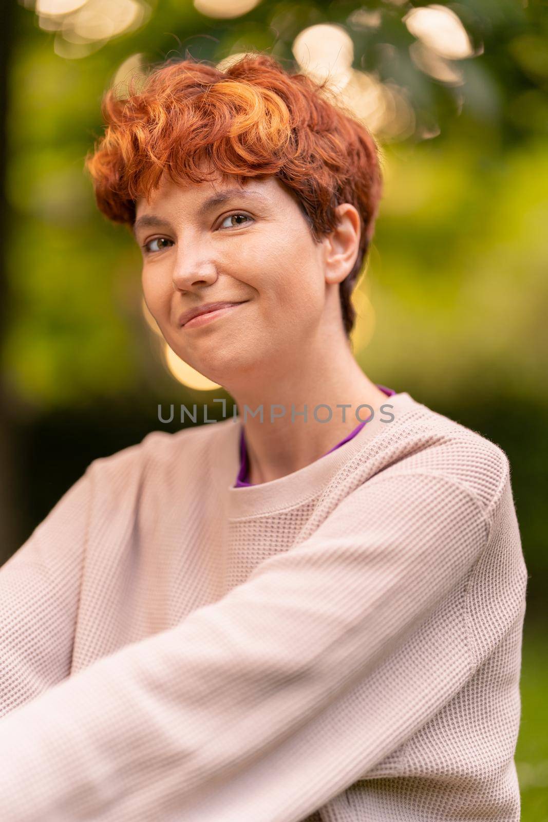 Glad lesbian female in casual sweatshirt with short ginger hair smiling and looking at camera on blurred background of summer park