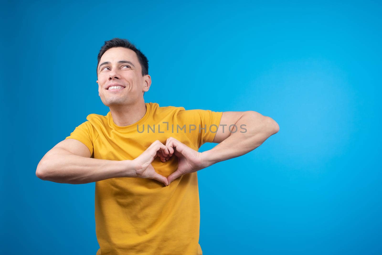 Happy male model in yellow t shirt making heart gesture and looking up with toothy smile on blue background