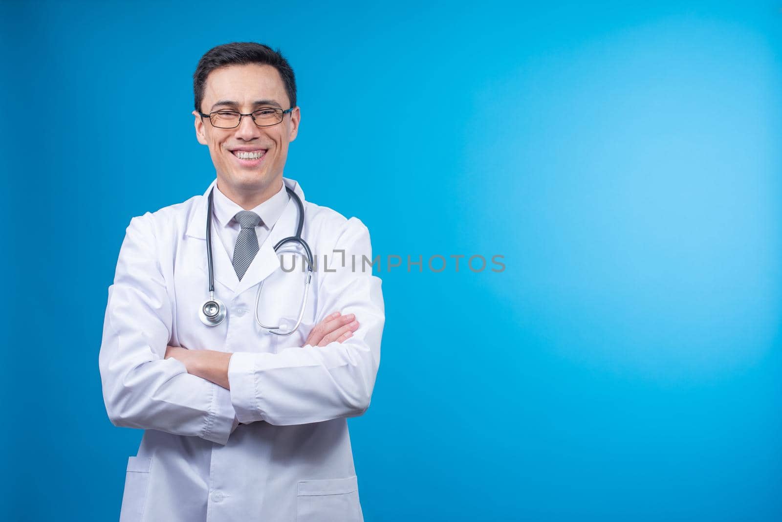 Glad doctor in white robe with stethoscope looking at camera with folded arms against blue background in studio