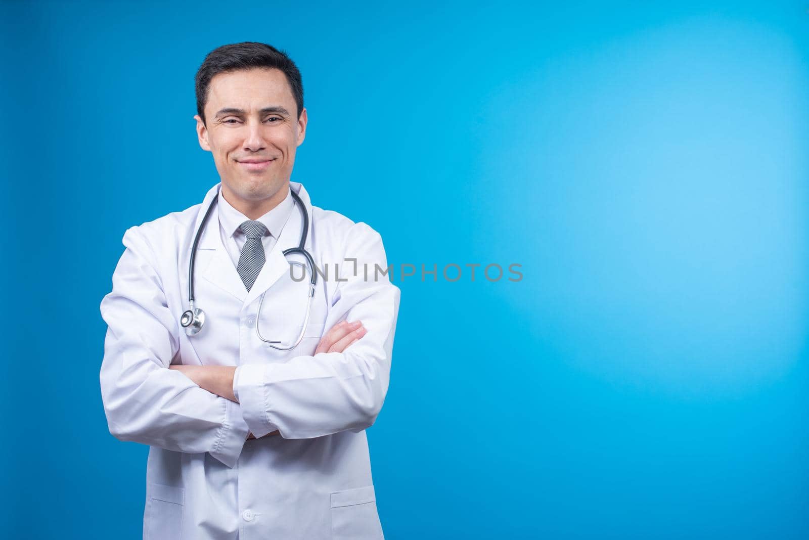 Pleasant doctor in medical uniform standing on blue background and looking at camera delightfully with stethoscope on neck