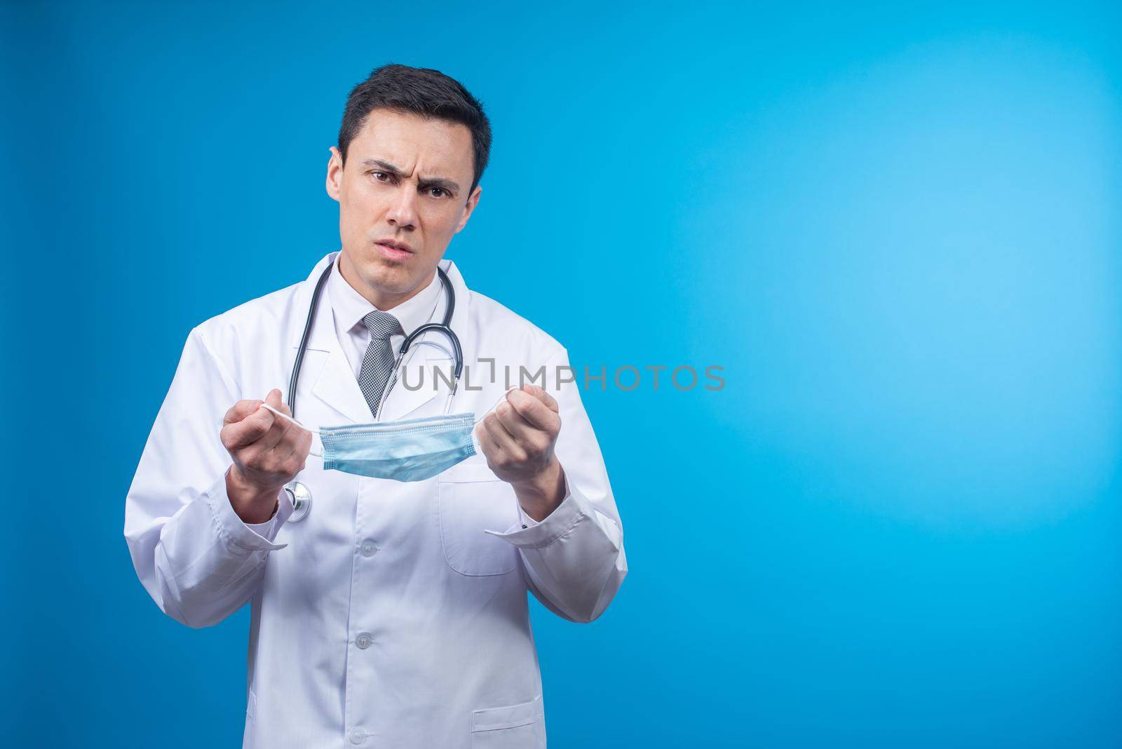 Physician in white robe with stethoscope frowning and looking at camera on blue background while holding mask