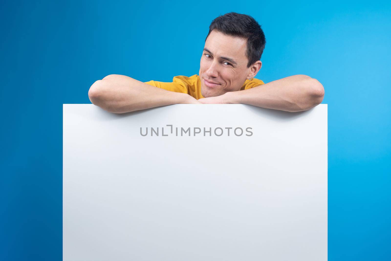 Smiling male model leaning on empty placard for advertisement and looking at camera with tricky face on blue background