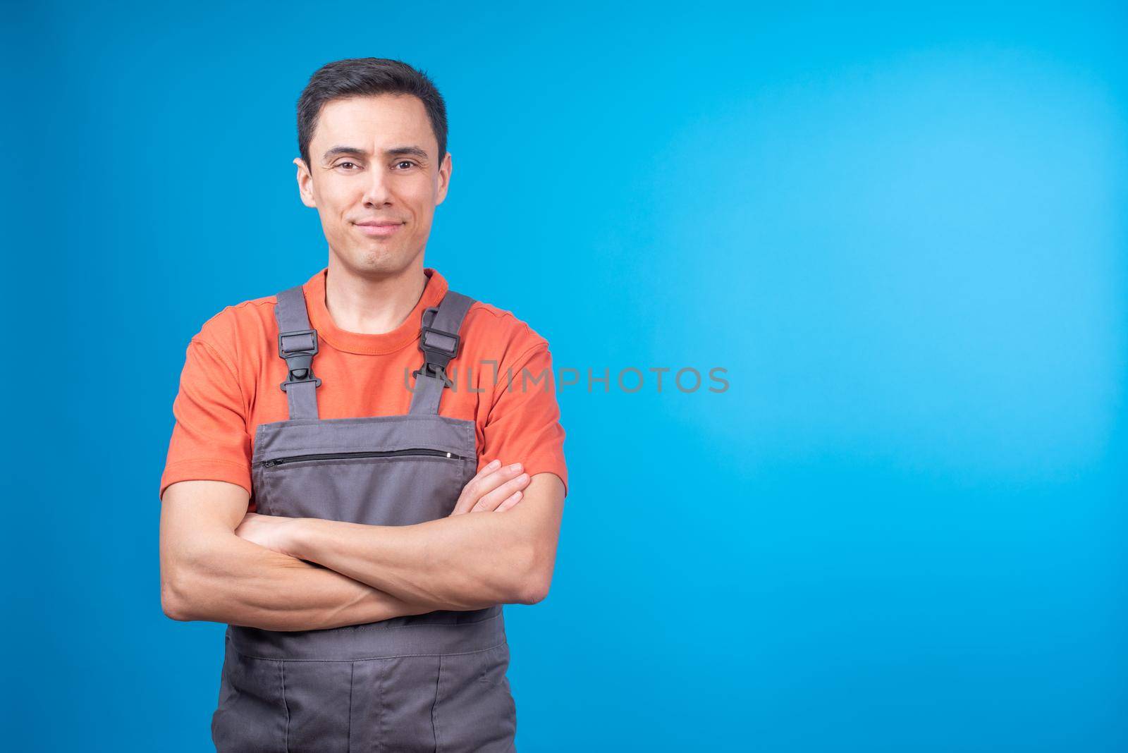 Self assured male contractor in workwear crossing arms and looking at camera against blue background