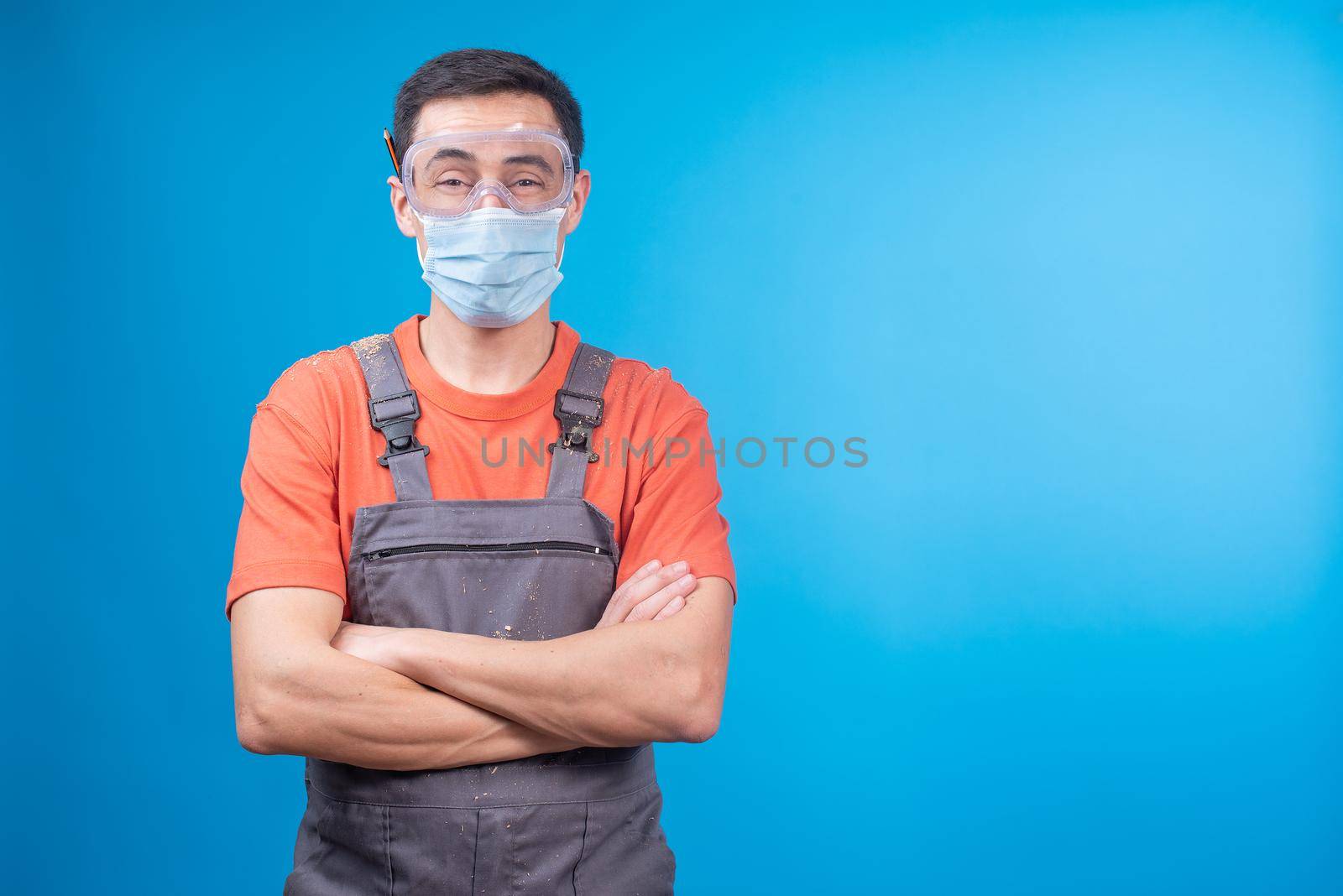 Self assured craftsman in uniform with protective goggles and face mask crossing arms and looking at camera against blue background during epidemic