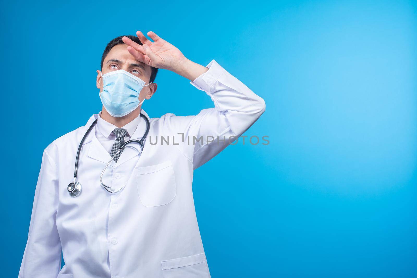 Tired physician in protective mask with stethoscope on neck putting hand on forehead against blue background