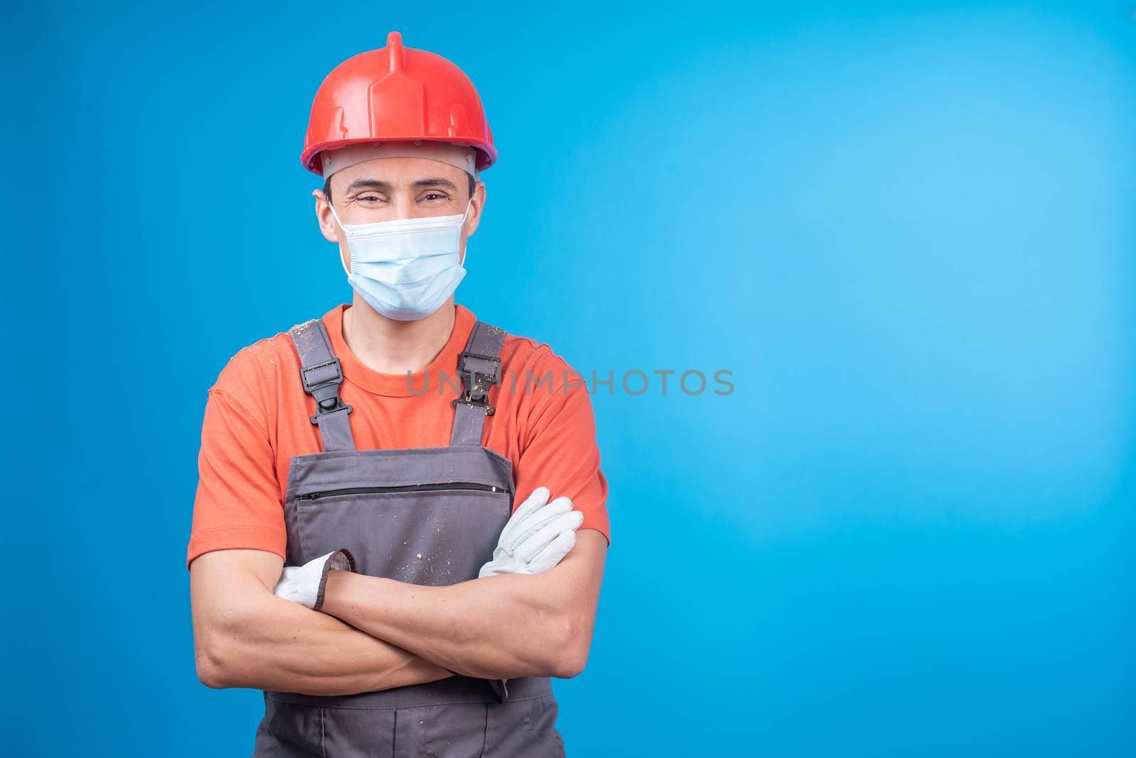 Confident male contractor in workwear with gloves, hardhat and face mask crossing arms and looking at camera while standing against blue background during epidemic