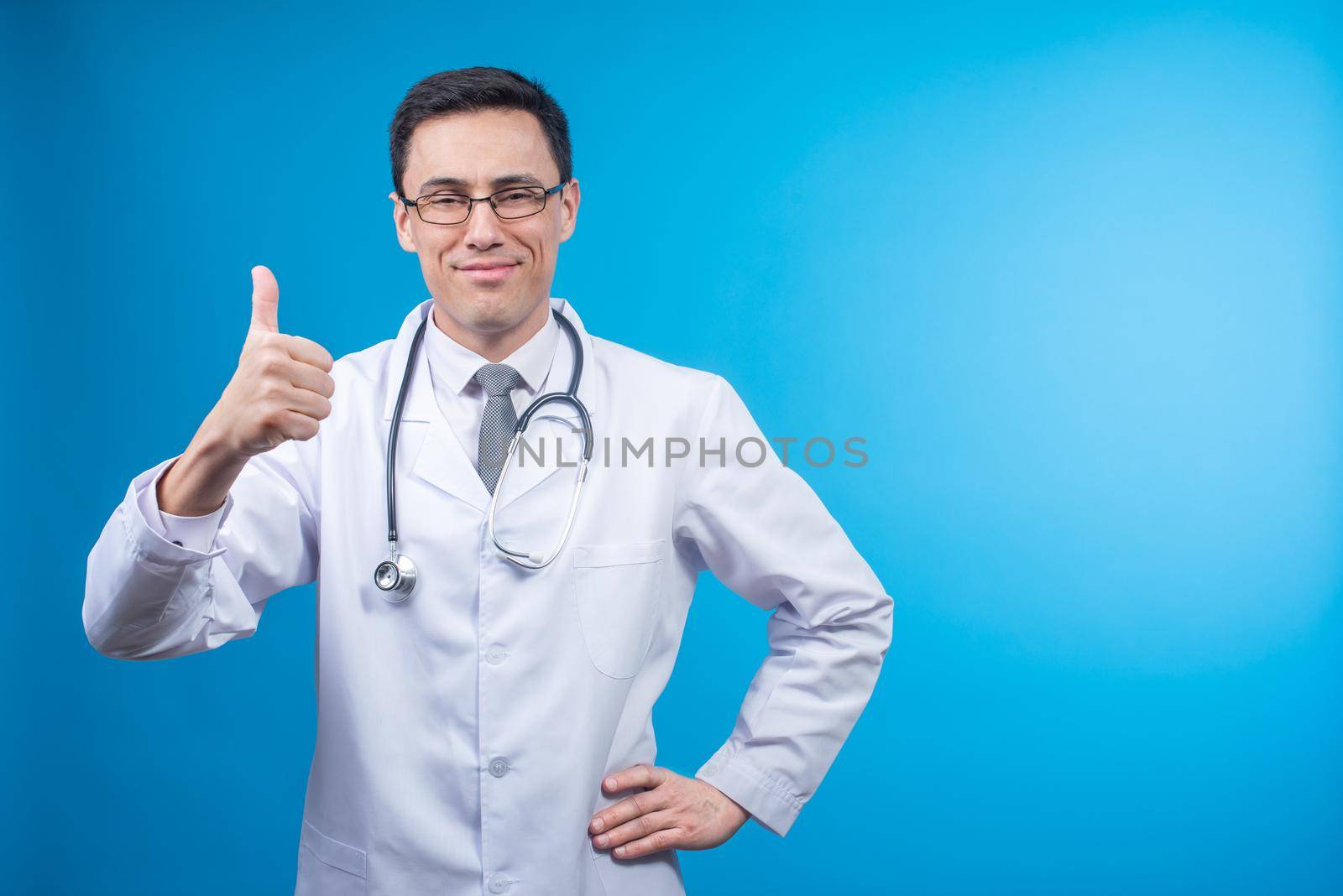 Satisfied male physician in white robe and stethoscope smiling and showing approve gesture against blue backdrop