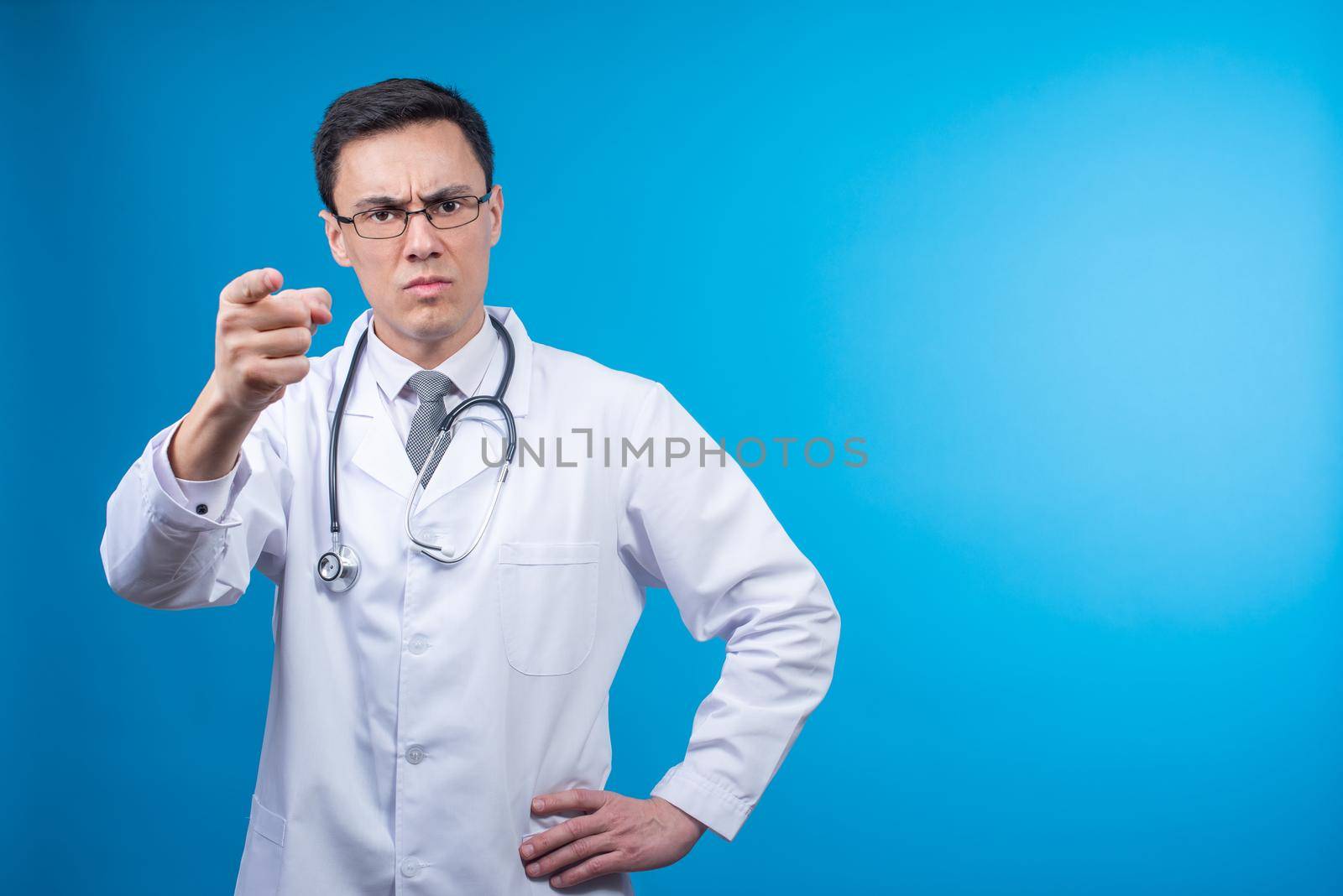 Isolated strict physician in white uniform and stethoscope on neck pointing at camera while standing against blue backdrop in studio