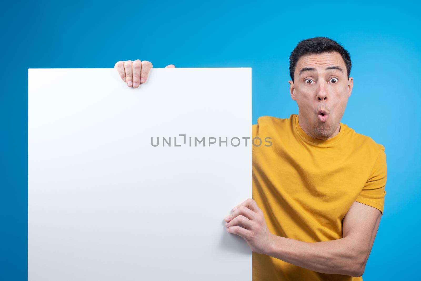 Startled guy in yellow t shirt looking at camera against blue background while demonstrating white sheet of paper