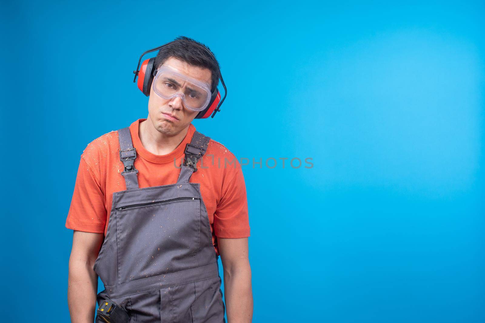 Unhappy craftsman in uniform with protective goggles and earmuffs pouting lips and looking at camera while being disappointed in work against blue background